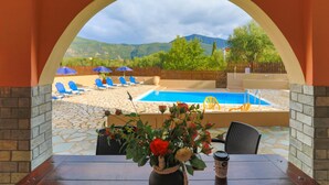 An outdoor seating area with a pool view, a table with a coffee cup, and blue umbrellas. - Villa Kristefi in Kato Korakiana (Corfu)
