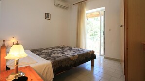 A bedroom with a bed, a bedside lamp, a small table, a window with curtains, and an air conditioning unit. - Alexandra Apartment in Katavolos (Corfu)