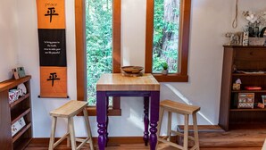 A room with a wooden table, two purple stools, a hanging banner with Chinese characters, and a window with a view of greenery. - Charming Tiny House nestled in the redwoods, less than a mile from the beach. (Fort Bragg)
