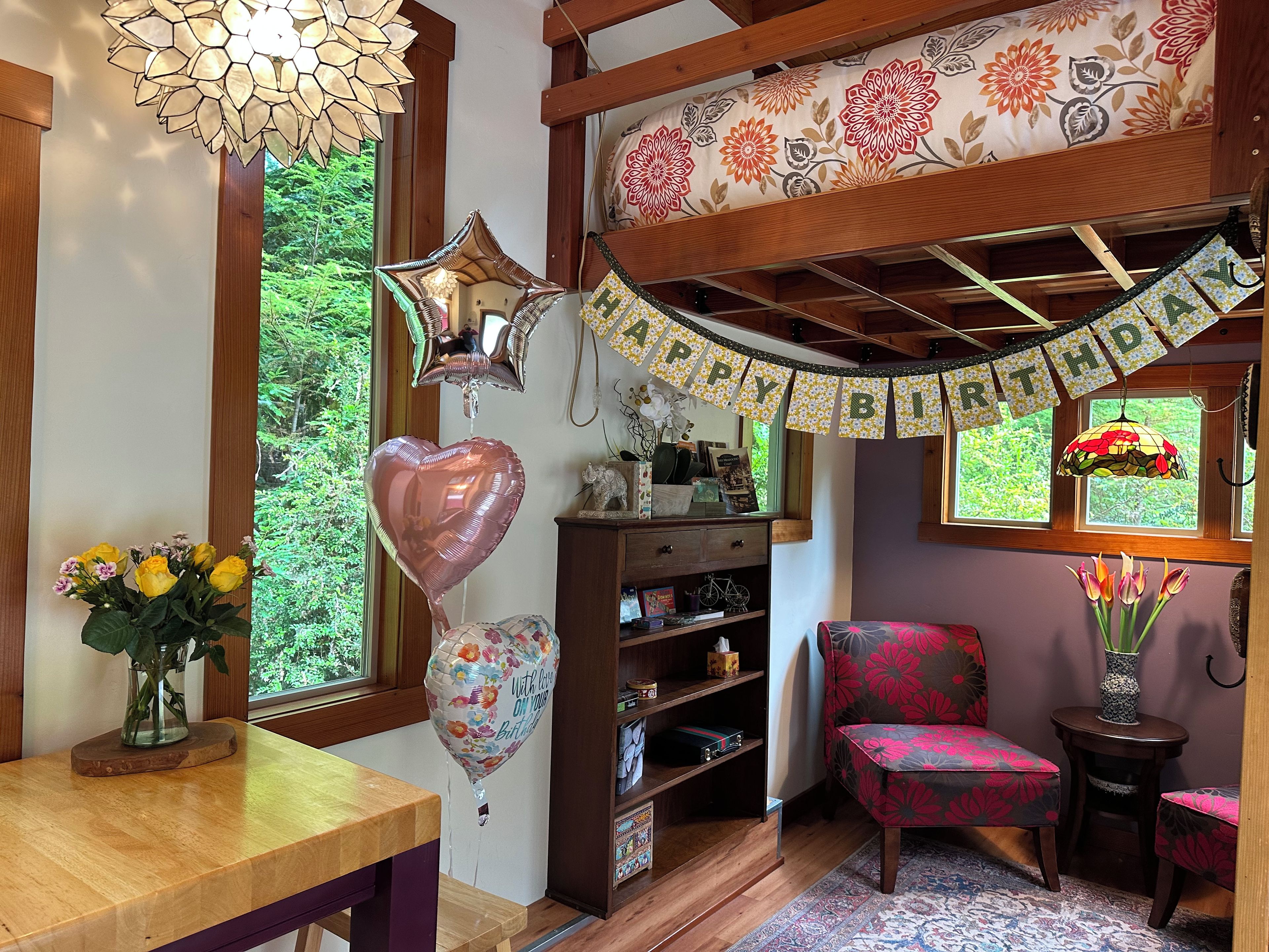 Una habitación acogedora con techo de madera, una estantería, un sillón estampado con motivos florales y una ventana con vistas a la vegetación.