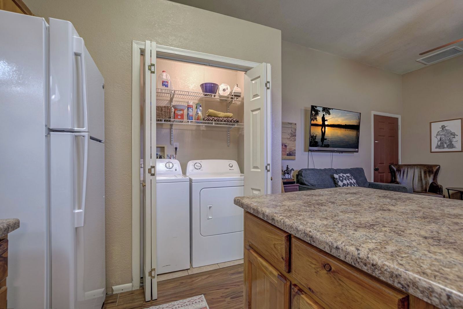 A compact kitchen with a refrigerator, washing machine, and dryer, a granite countertop, and a dining area with a couch and a painting.
