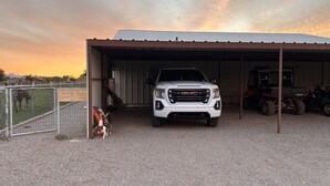 A white GMC truck parked inside a covered garage with a gravel floor, a fenced area with a zebra visible in the background, and a sign reading "Guest House".