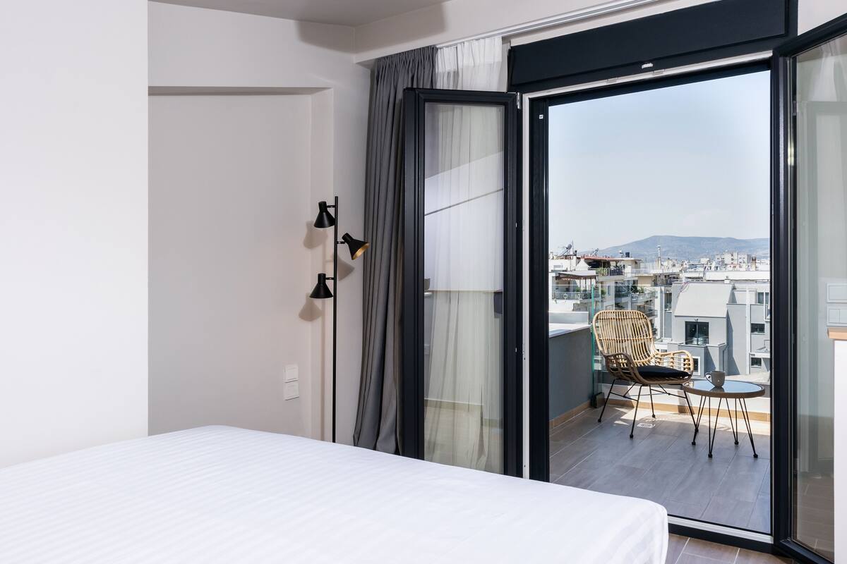 Habitación de hotel moderna con una cama grande, un balcón con una silla y una mesa, y vistas a la ciudad.