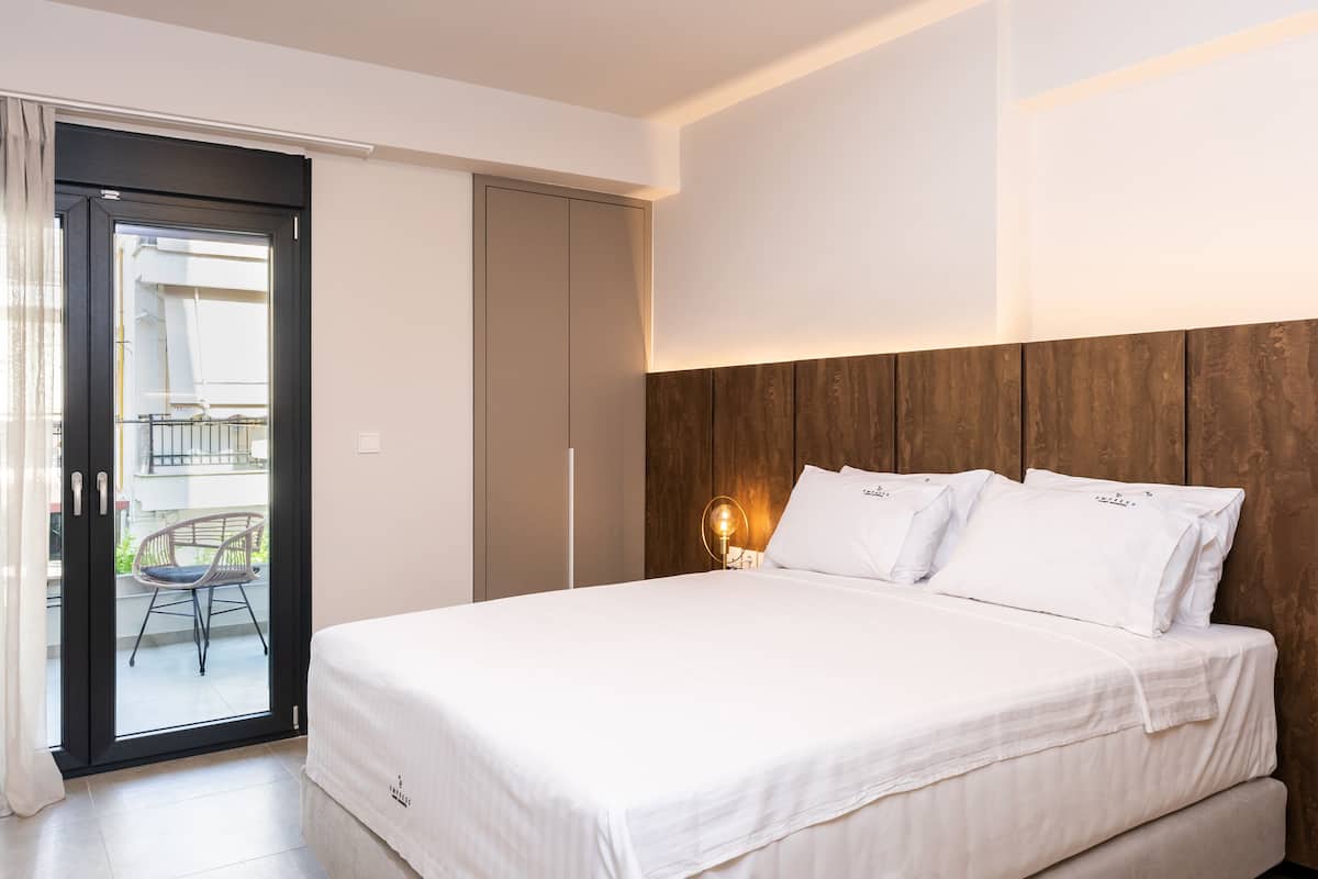 Un dormitorio con cama, cabecera de madera, mesita de noche con lámpara y puerta corrediza que da a un espacio exterior con una silla.