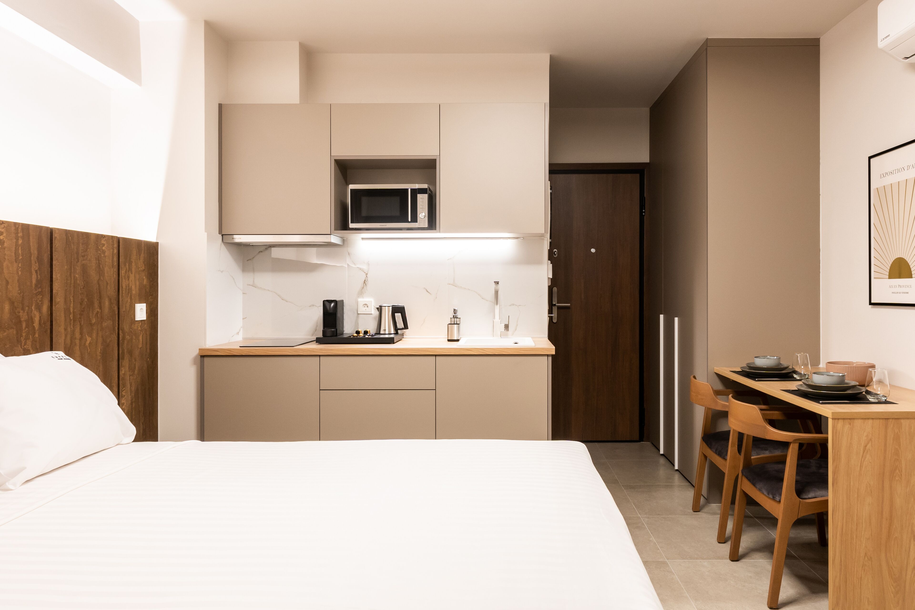 A compact hotel room with a bed, a kitchenette, a dining area, and a door leading to a bathroom.