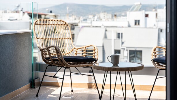 Un balcón con sillas de mimbre y una pequeña mesa, con vistas a un paisaje urbano que incluye edificios y montañas.