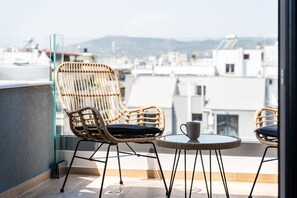 A balcony with wicker chairs and a small table, overlooking a cityscape with buildings and mountains. - Empress Luxury Residences (Volos)