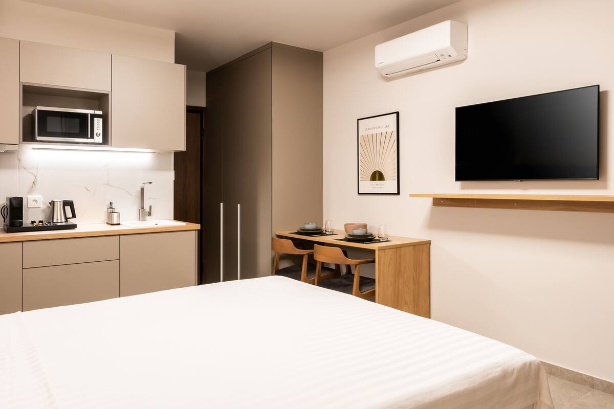 Una habitación de hotel moderna con cama, escritorio con dos sillas, microondas, cafetera y televisor de pantalla plana montado en la pared.