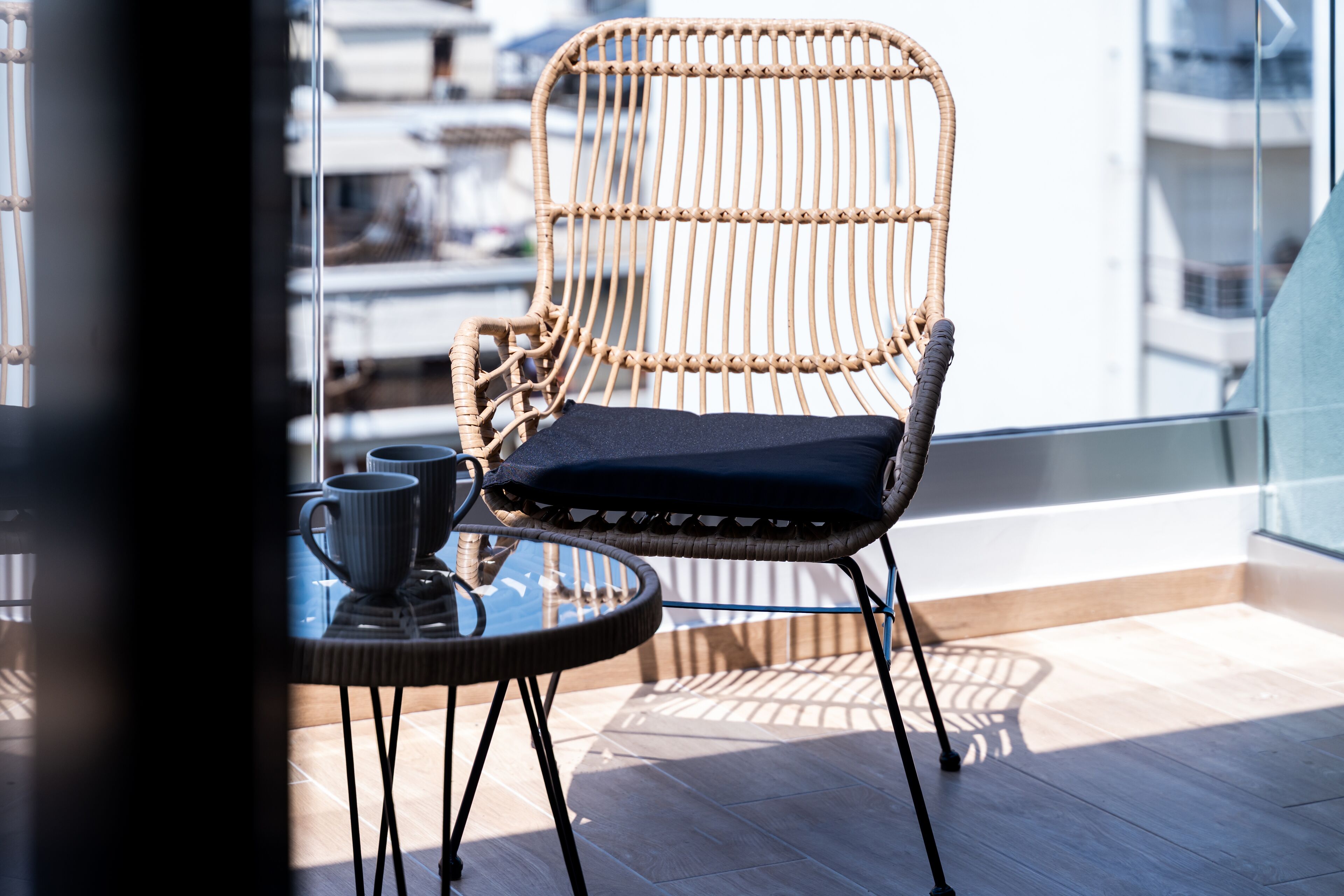 A wicker chair with a cushion, a glass table with two cups, and a view of a building through a window.
