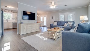 Un vaste salon comprenant un grand canapé, une table basse et un téléviseur fixé au mur. On y trouve également un coin cuisine avec des meubles blancs et une table à manger.