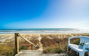Eine Holztreppe führt zu einem Strand, mit einem weißen Kunststoffstuhl und einem Zaun.