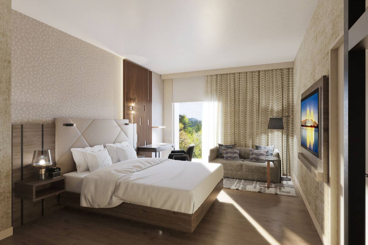 A modern hotel room with a large bed, a desk, a chair, and a sofa. There is a window with a view of greenery and a painting on the wall.