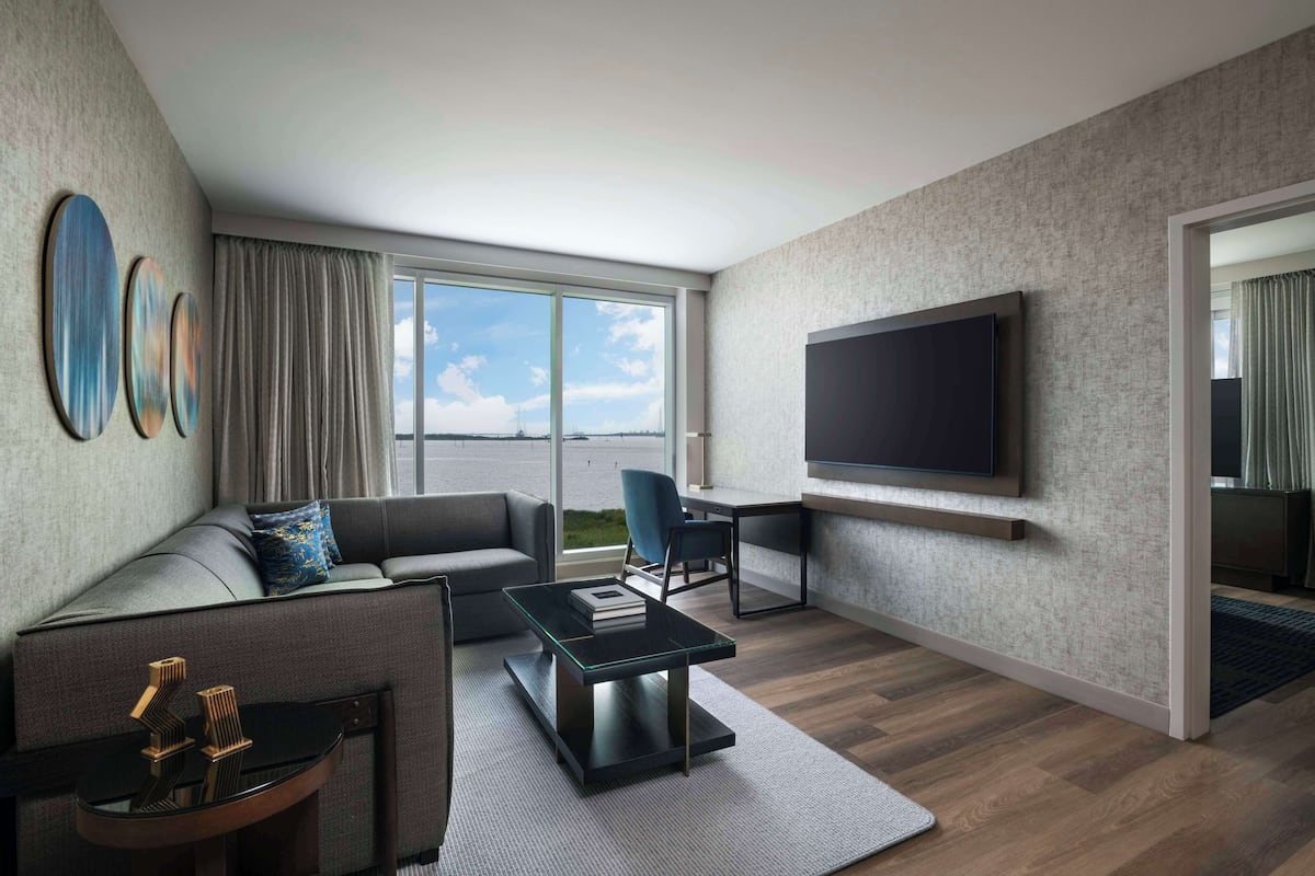 A modern living room with a sofa, a coffee table, a TV mounted on the wall, and a view of the outdoors through large windows.