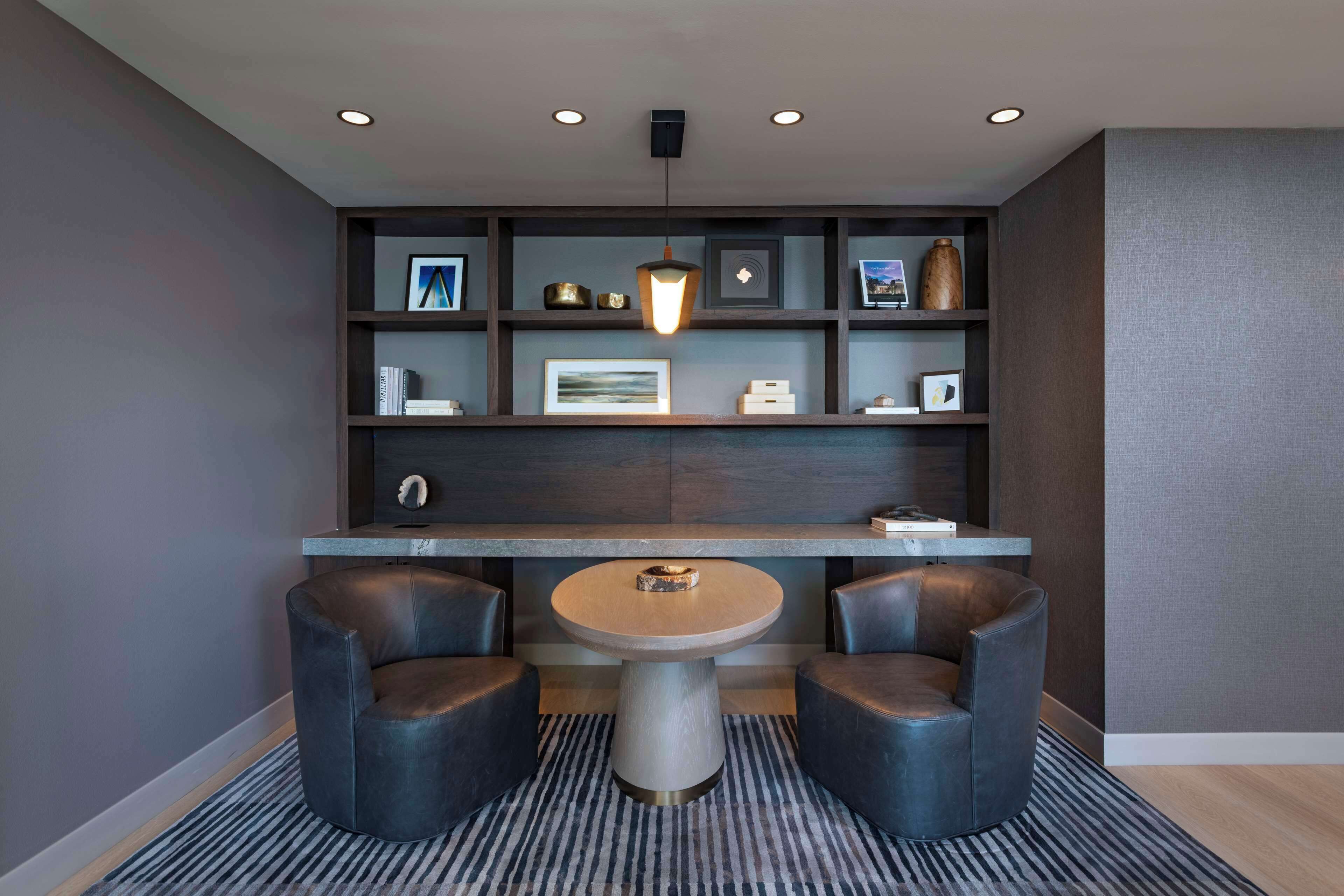 A modern room with a round table and two chairs, a bookshelf with various items, and a hanging light fixture.