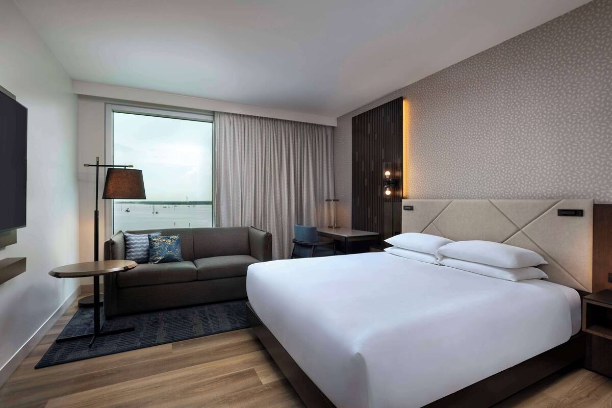 A modern hotel room with a large bed, a sofa, a small table, and a desk.