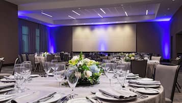 Un salón de banquetes con mesas redondas dispuestas para cenar, una pantalla grande al frente e iluminación ambiental.