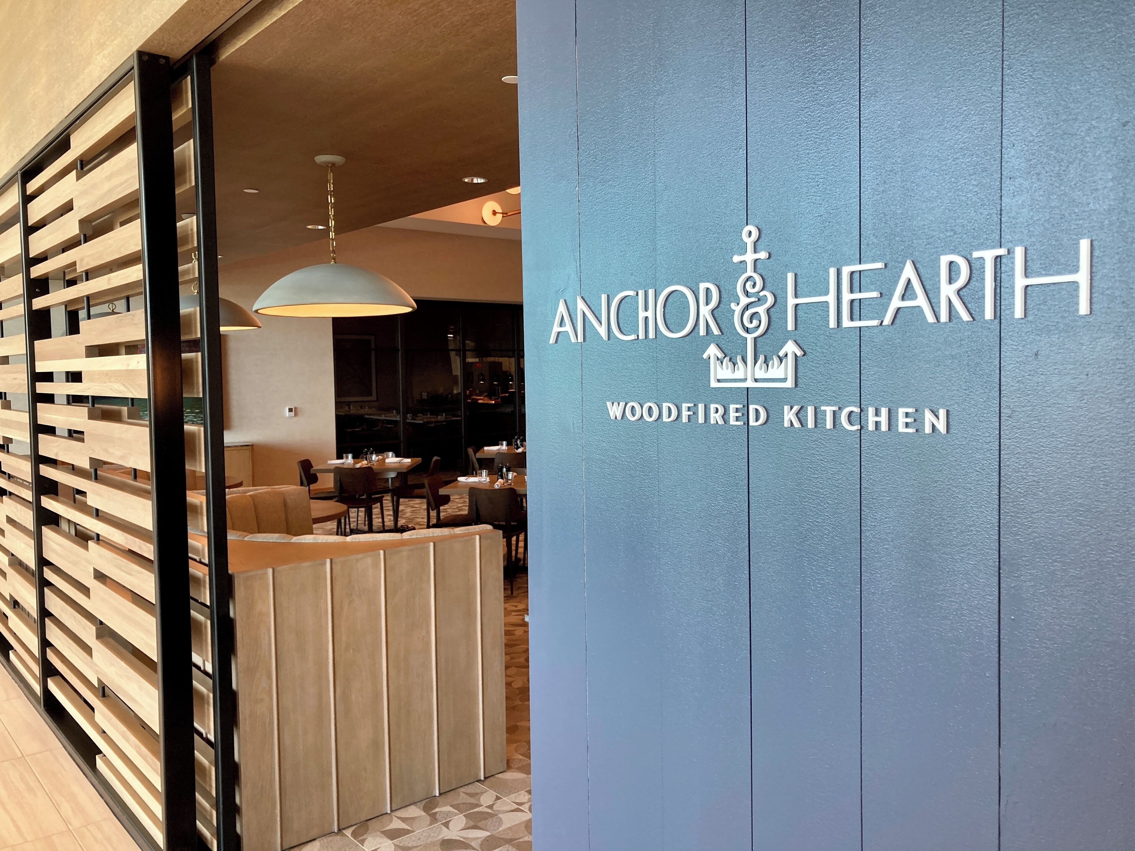A restaurant interior with a modern design, featuring a wooden partition and a sign that reads "Anchor & Hearth Woodfired Kitchen".
