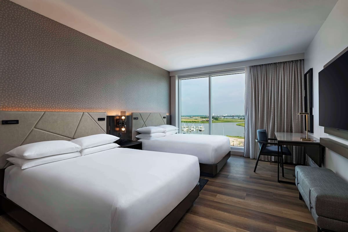 A hotel room with two beds, a desk, a chair, a TV, and a view of a marina.