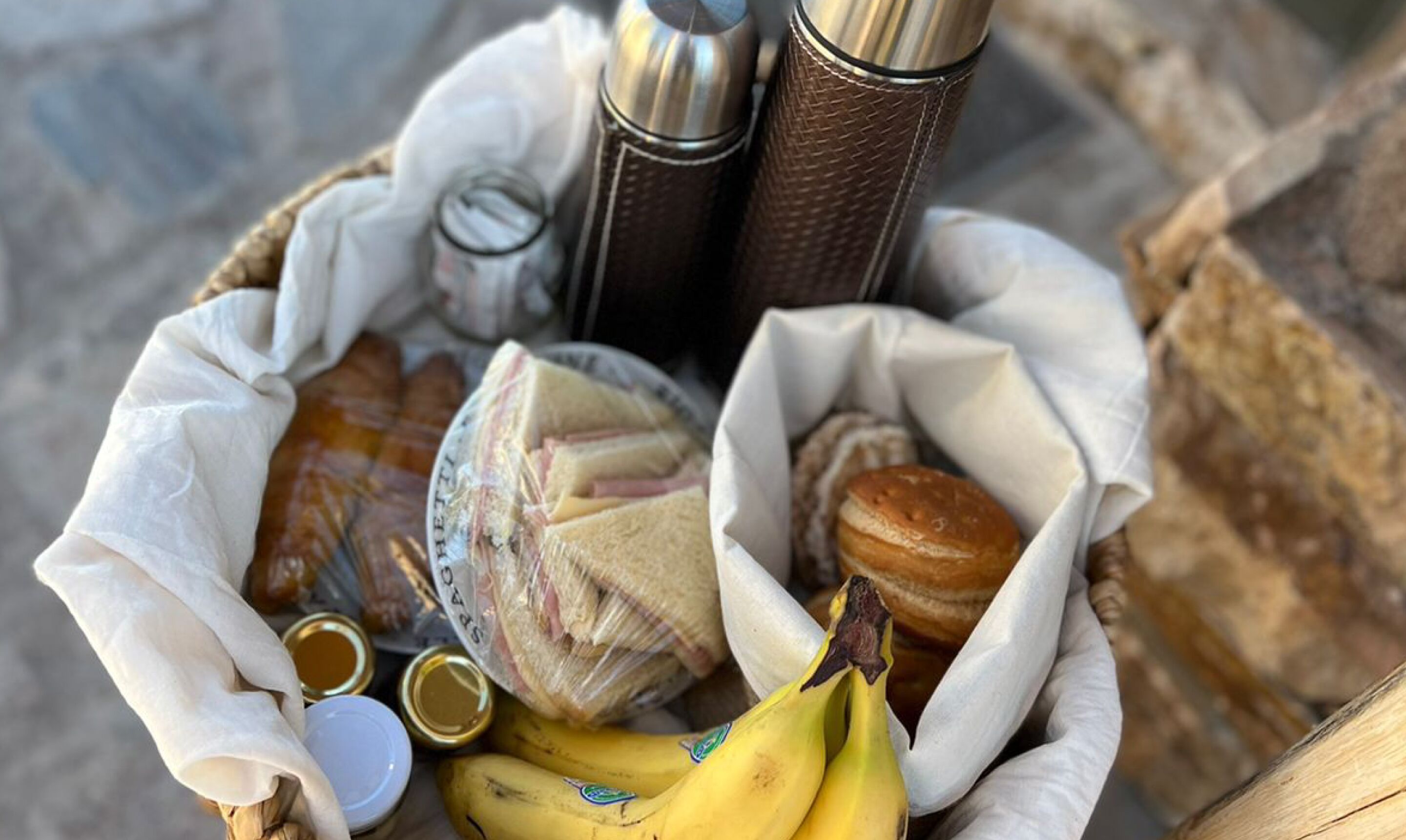 A basket containing sandwiches, a baguette, bananas, condiments, and a thermos.