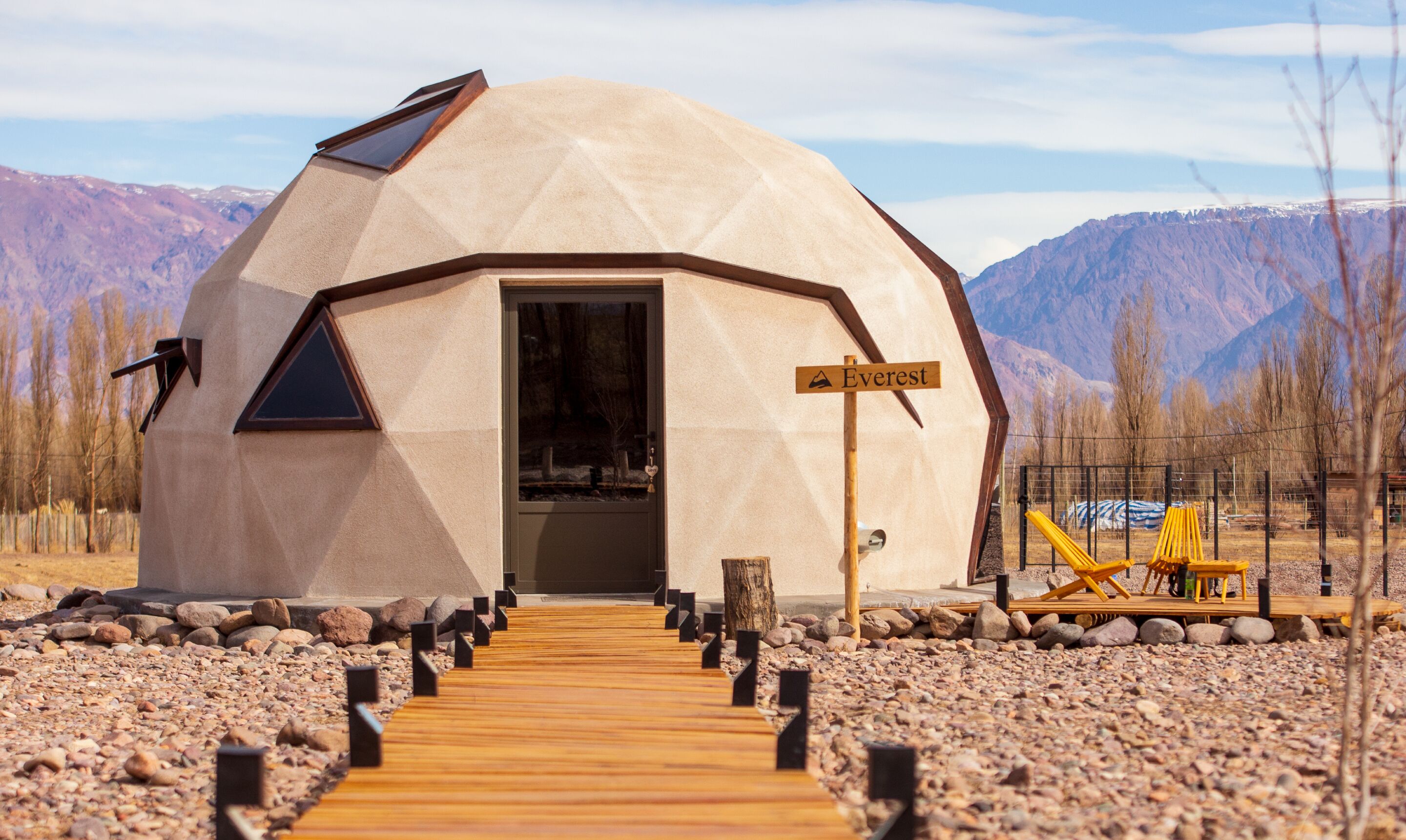 A geodesic dome structure with a wooden deck leading to the entrance, surrounded by a mountainous landscape.