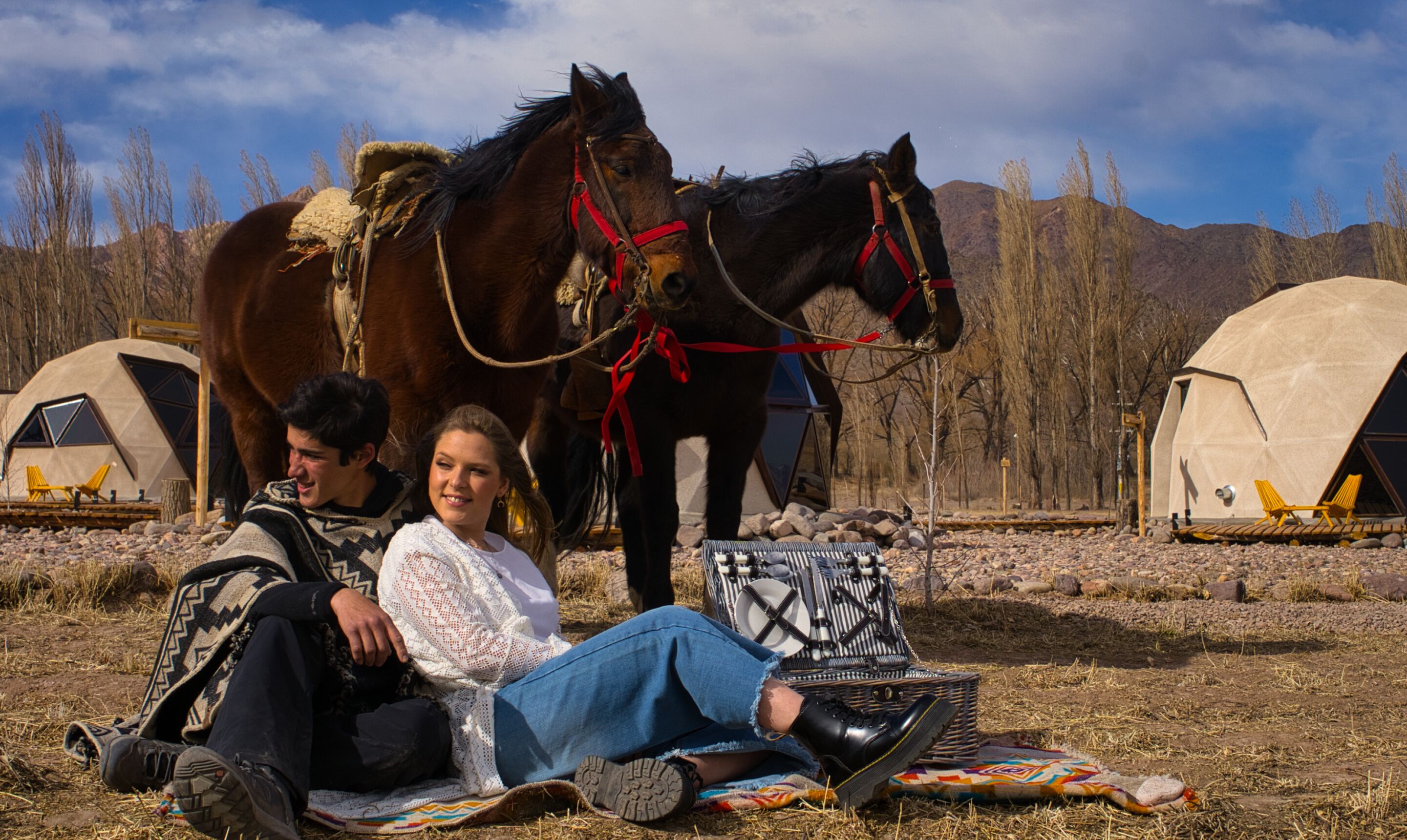 A couple sitting on the ground with a horse and two yurts in a natural setting.