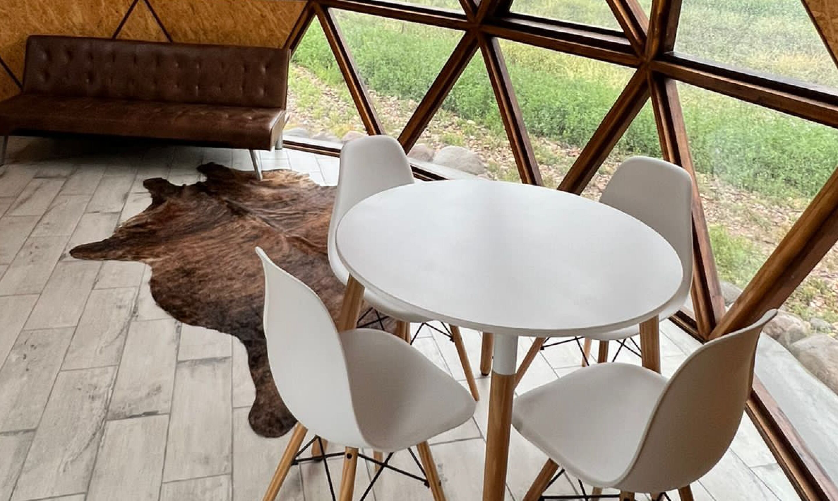 Una habitación con una mesa redonda y cuatro sillas, un sofá de cuero marrón y un ventanal con vistas al verdor.