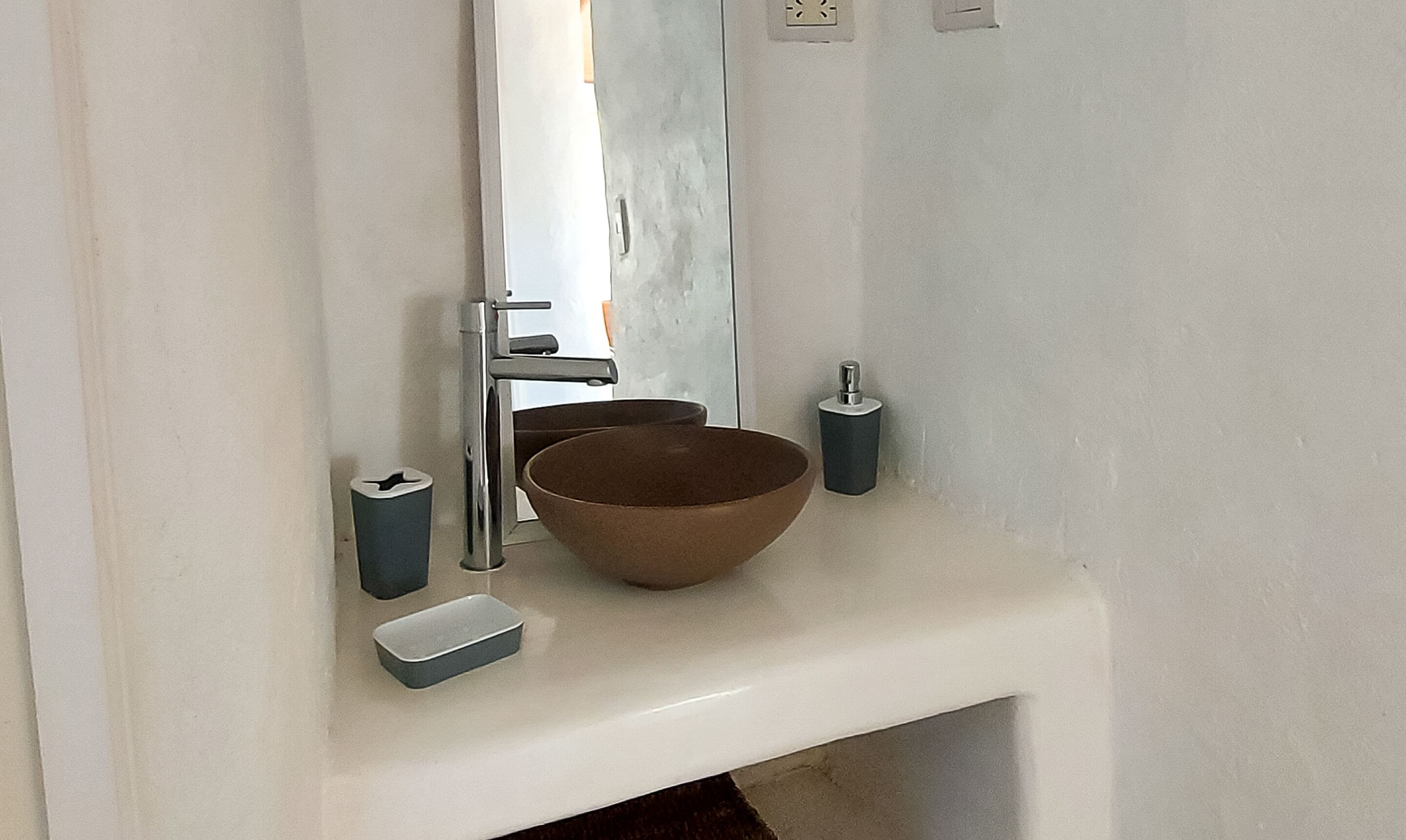 Un baño con un lavamanos blanco, un cuenco marrón, un dispensador de jabón y un bote de basura.