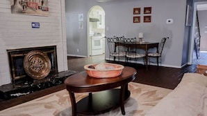 Ein Wohnzimmer mit Kamin, eine Essecke mit Tisch und Stühlen sowie eine durch einen Bogen sichtbare Küche.