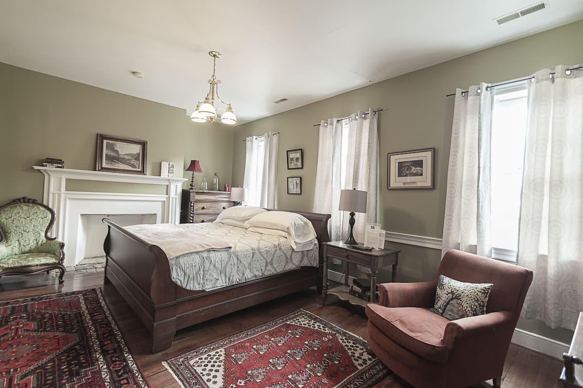 A bedroom with a bed, a chair, a nightstand, a lamp, a fireplace, and a window with curtains.