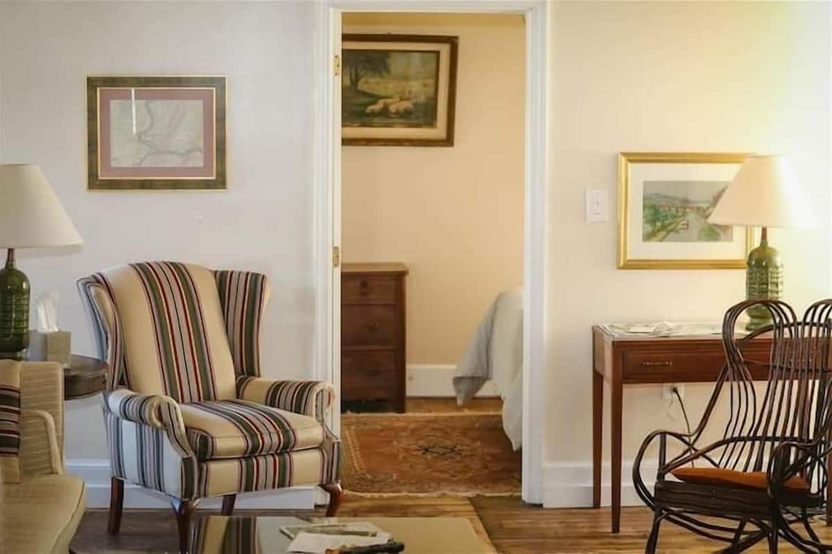 A room with a striped armchair, a wooden desk, and a chair with a cushion. There are framed pictures on the walls and a lamp on the desk.