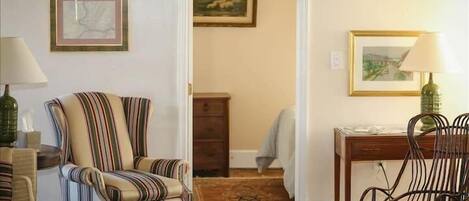 Une pièce avec un fauteuil à rayures, un bureau en bois et une chaise avec un coussin. Des tableaux encadrés ornent les murs et une lampe se trouve sur le bureau.