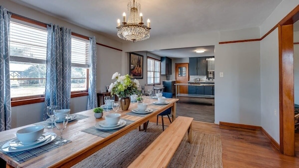 A dining area with a long wooden table set for four, a chandelier, and a view of the kitchen in the background. - Totally Turquoise (Savannah)