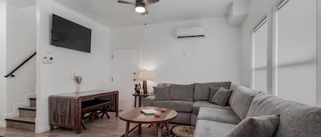 A modern living room with a grey sectional sofa, a wooden coffee table, a red patterned rug, a ceiling fan, a flat-screen TV mounted on the wall, and a staircase leading to another level.