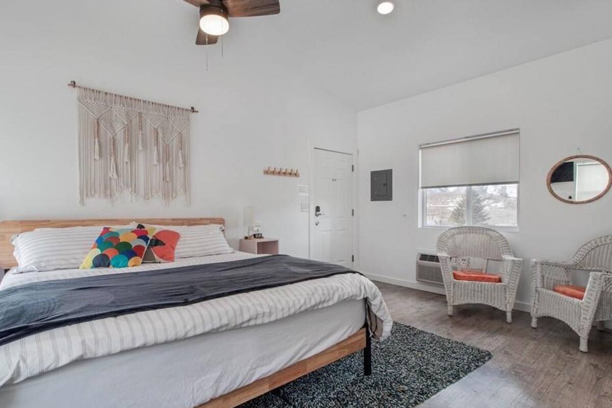 Une chambre avec un lit, une décoration murale en macramé, des chaises en osier et une grande fenêtre.