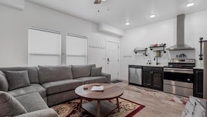 Un salon moderne avec un canapé gris, une table basse ronde en bois et un coin cuisine doté d’appareils électroménagers en acier inoxydable.
