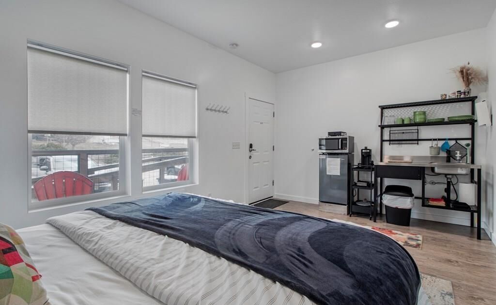 A modern bedroom with a large bed, a desk with a computer, a microwave, and a small kitchenette.