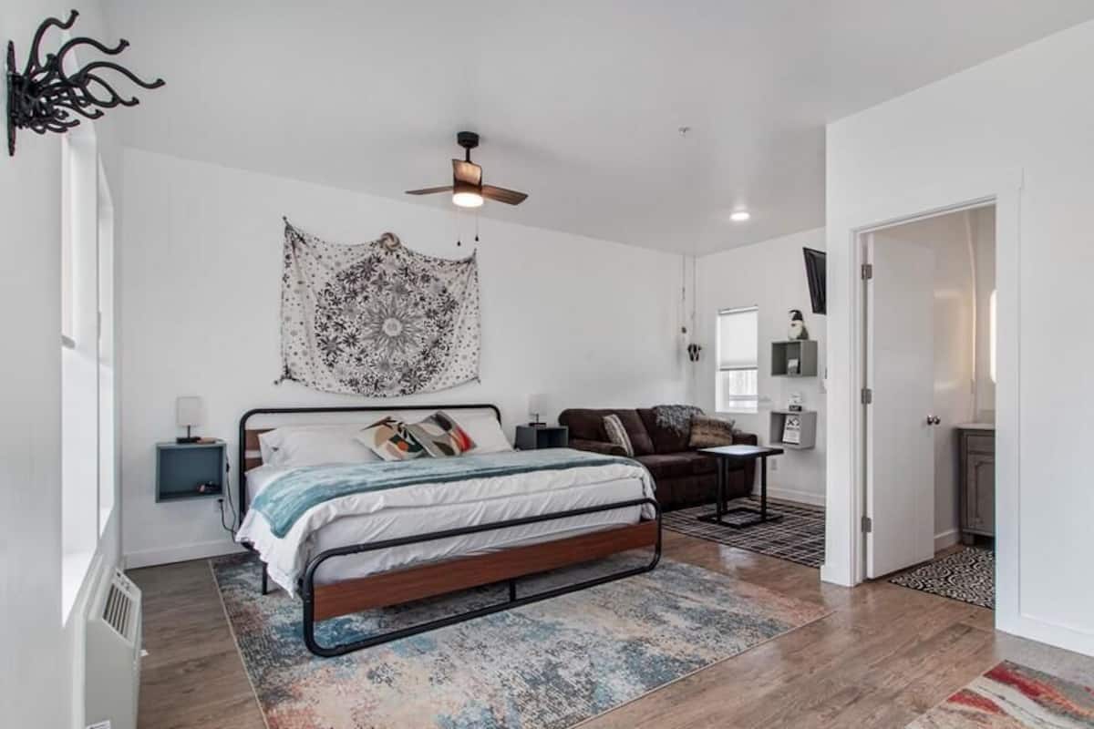 Une chambre à coucher comprenant un lit, un ventilateur de plafond, une tapisserie murale, une table de chevet, une commode et une porte donnant sur une autre pièce.