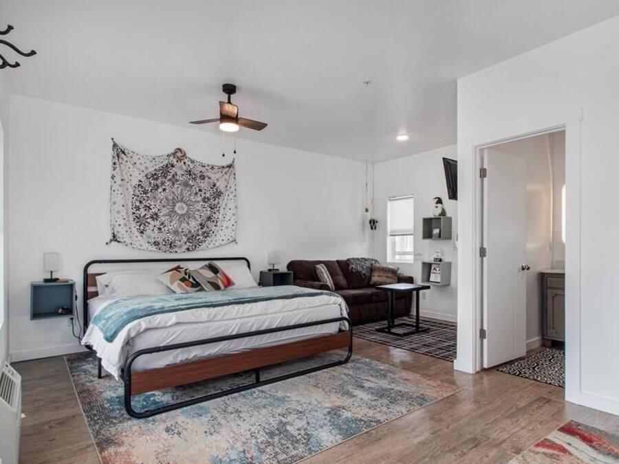 Un dormitorio con cama, ventilador de techo, tapiz, mesita de noche, cómoda y una puerta que da a otra habitación.