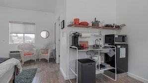 Une petite chambre comprenant un lit, deux fauteuils en osier, une petite table, un coin cuisine équipé d’un four à micro-ondes et d’une machine à café, ainsi qu’une poubelle.