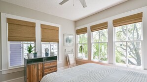 A bedroom with a large bed, wooden dresser, and a vanity with a mirror. There are large windows with blinds and a view of greenery outside. - Blue Mermaid! Beautiful home 4 blocks from the beach! (Galveston)