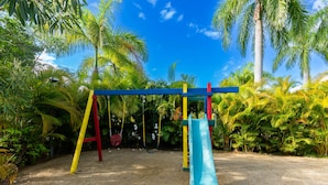 Ein Spielplatz mit Rutsche und Schaukeln, umgeben von Palmen und viel Grün.