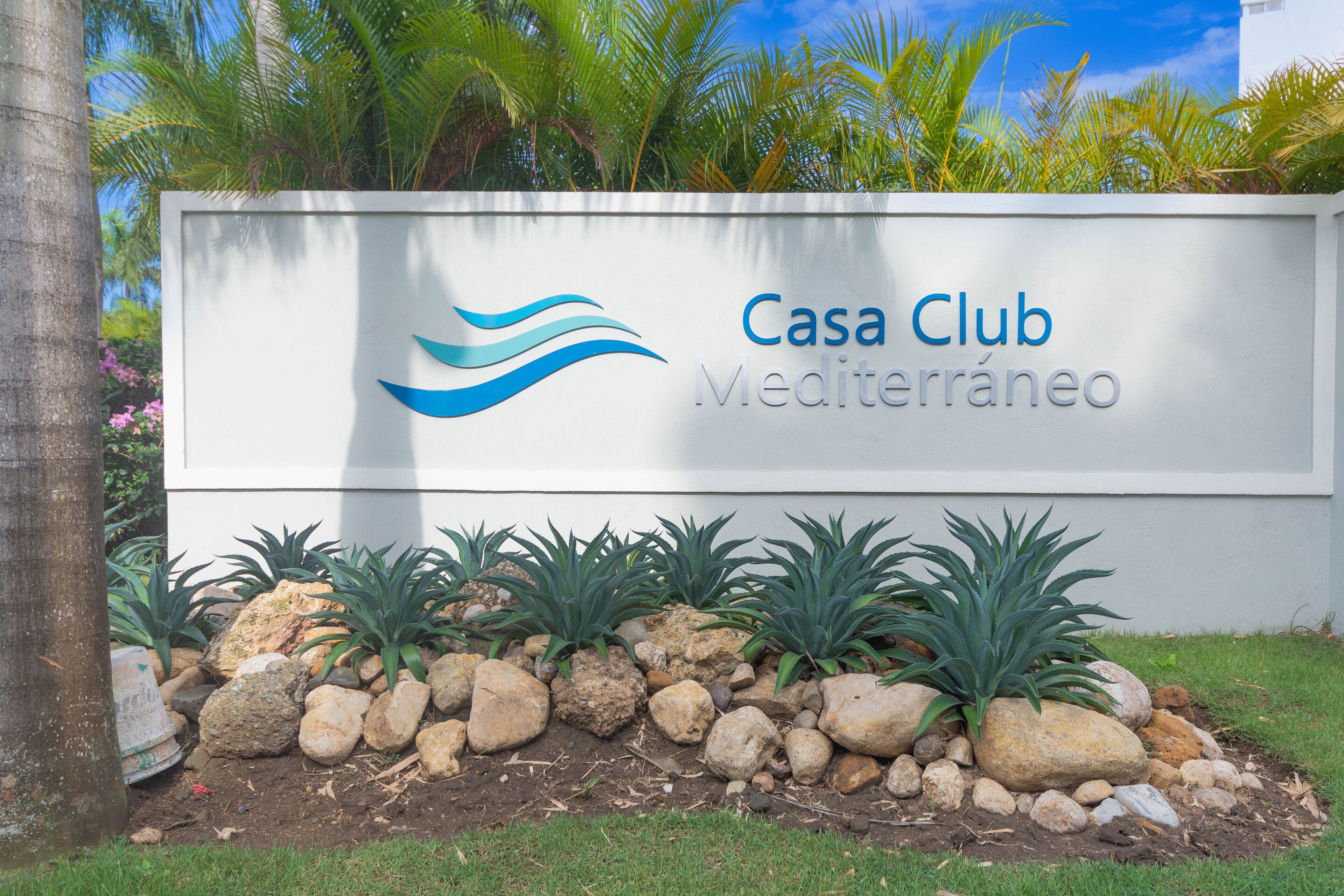 A sign for Casa Club Mediterraneo with a landscaped garden featuring rocks and plants.