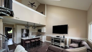 A spacious living area with a high ceiling, a kitchen with a central island, a large flat-screen TV mounted on the wall, and a comfortable seating area with a sofa and a chair.