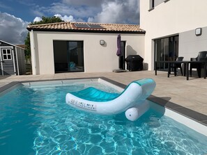 Une piscine avec un flotteur gonflable bleu, une terrasse avec des chaises et un bâtiment doté d’un toit en tuiles et de panneaux solaires.