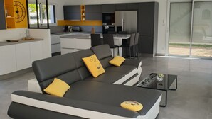 Una sala de estar moderna con un sofá grande en forma de L, un comedor y una cocina al fondo, y un ventanal corredizo que da a un espacio exterior.