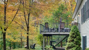 A covered outdoor deck with chairs and tables, surrounded by trees with autumn foliage.