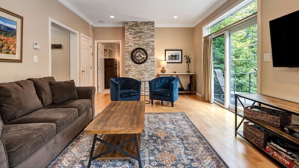 A living room with a brown sofa, two blue armchairs, a wooden coffee table, a patterned rug, a stone accent wall, a mounted clock, and a large window with a view of greenery.