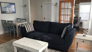 Un salón con un sofá azul oscuro, una mesa de centro blanca y un cojín a rayas. En la pared hay un cuadro, una puerta con un panel de cristal y una lámpara sobre una mesita.
