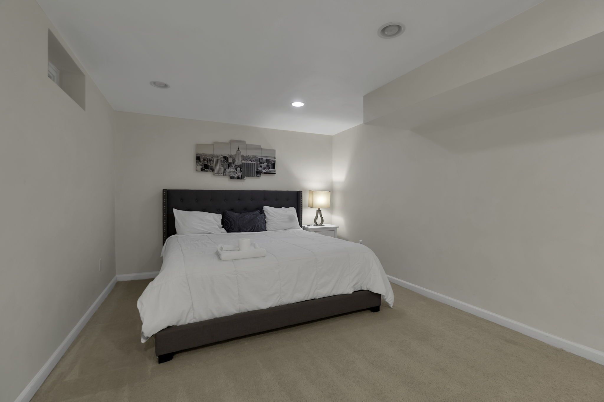 A neatly made bed with a dark headboard, a bedside lamp, and a cityscape artwork on the wall.