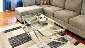 Un salon comprenant un canapé d’angle, une table basse en verre, un rideau bleu, un téléviseur fixé au mur et un grand tableau abstrait.